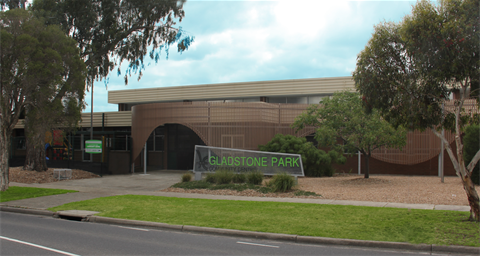 Photograph of Gladstone Park Community Centre building with 70's architectural design, featuring a large sign with green text on a clear background in front of the entrance. Surrounding elements include trees and a sparce maintained garden