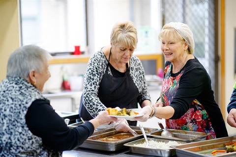 Two ladies serve lunch to an older lady