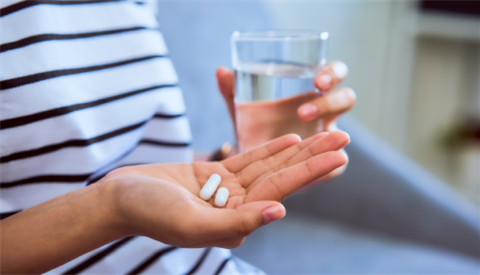 Woman holding white pill on hand and drinking water in glass on sofa in house, feels like sick.